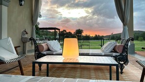 Eine Terrasse mit Tisch, zwei Sofas, einer Lampe und Blick auf die Landschaft bei Sonnenuntergang.