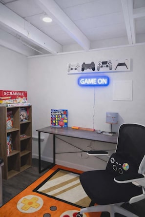 Ein Zimmer mit Schreibtisch, Stuhl, einer Spieleauswahl und einem Neon-Schild mit der Aufschrift 'GAME ON'.