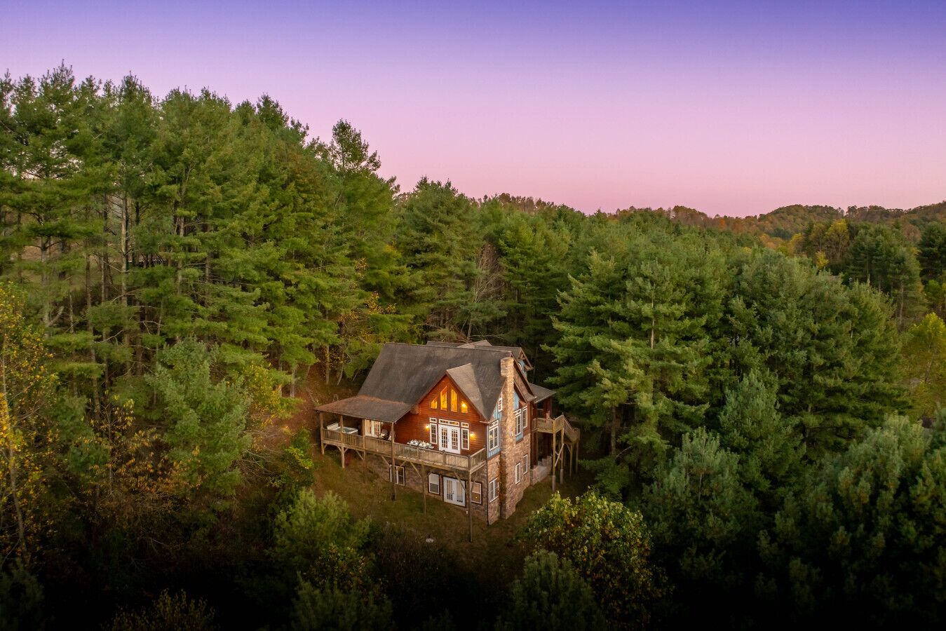 A cabin surrounded by dense forest, featuring a prominent front porch and a large chimney.