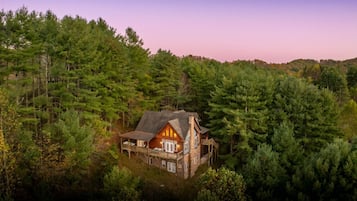 A cabin surrounded by dense forest, featuring a prominent front porch and a large chimney.
