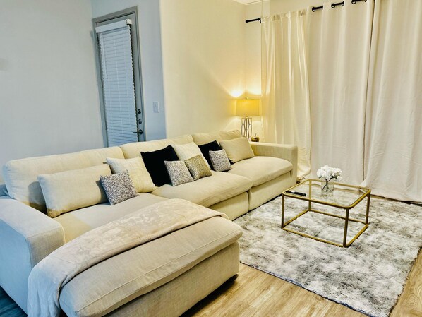 Un salon avec un canapé d’angle beige, une table basse en verre, un tapis moelleux, une lampe et une porte avec des stores.