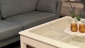 A living room with a grey sofa, a glass coffee table with vases, a bookshelf, a floor lamp, and a window with a view.