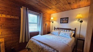 Quarto aconchegante com teto de madeira, cama com roupa de cama floral, duas luminárias de parede e janela com persianas.
