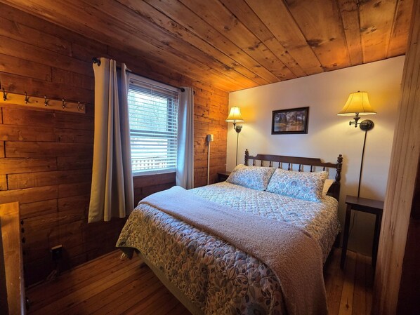A cozy bedroom with a wooden ceiling, a bed with floral bedding, two wall-mounted lamps, and a window with blinds. - Mountain View Log Cabin - Cozy cabin in the Blue Ridge Mountains (Stanley)