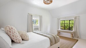 Un dormitorio con una cama grande, dos ventanas con cortinas, un banco y una lámpara en el techo.
