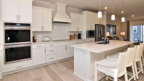 A modern kitchen with white cabinetry, stainless steel appliances, a central island with bar stools, pendant lighting, and a view of the living area. - BRAND NEW North OC 3BR + Loft Sleeps 12, 5 baths Walk to Pool & Beach (Ocean City)