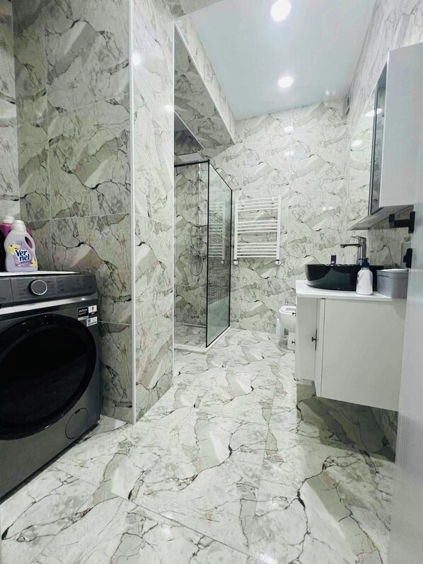A bathroom with a marble floor, a glass shower enclosure, a washing machine, and a sink with a mirror above it.