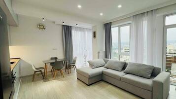 A modern living room with a grey sectional sofa, a dining table with chairs, and a view of the city through large windows.