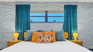 Ein Bett mit weißer Bettdecke, zwei orangefarbenen Kissen und einem gemusterten Kissen. Zwei Nachttische mit Lampen stehen daneben. Im Raum befindet sich ein Fenster mit blauen Vorhängen und Blick auf Himmel und Häuser.