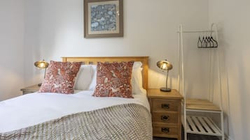 A neatly made bed with patterned pillows, a bedside table with a lamp, a small wooden nightstand, and a wall-mounted coat rack.