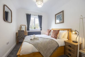A neatly arranged bedroom with a wooden bed, a bedside lamp, a nightstand, a chair, a window with curtains, a mirror, and a framed picture on the wall. - Beautiful 3-Bed Home with Hot Tub in Ross-on-Wye (Ross-on-Wye)