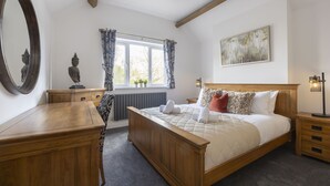 A bedroom with a wooden bed, a bedside table, a dresser, a window with curtains, a radiator, a painting on the wall, and a mirror.