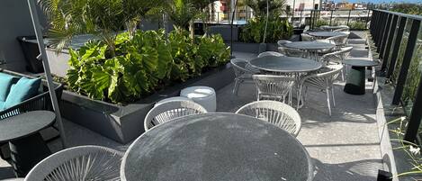 Eine Dachterrasse mit runden Tischen und Korbstühlen, umgeben von Grünpflanzen und mit Blick auf eine Stadtlandschaft.
