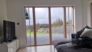 A modern living room with a flat-screen TV, a grey sofa, and a sliding glass door leading to an outdoor area. - A large 4 bed family home to make great memories in the West Coast of Ireland. (Westport)