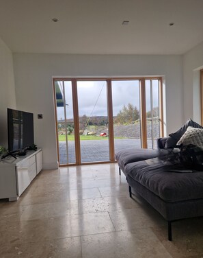 A modern living room with a flat-screen TV, a grey sofa, and a sliding glass door leading to an outdoor area. - A large 4 bed family home to make great memories in the West Coast of Ireland. (Westport)