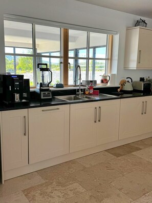 A modern kitchen with a large window, a coffee machine, a blender, and a kettle. - A large 4 bed family home to make great memories in the West Coast of Ireland. (Westport)