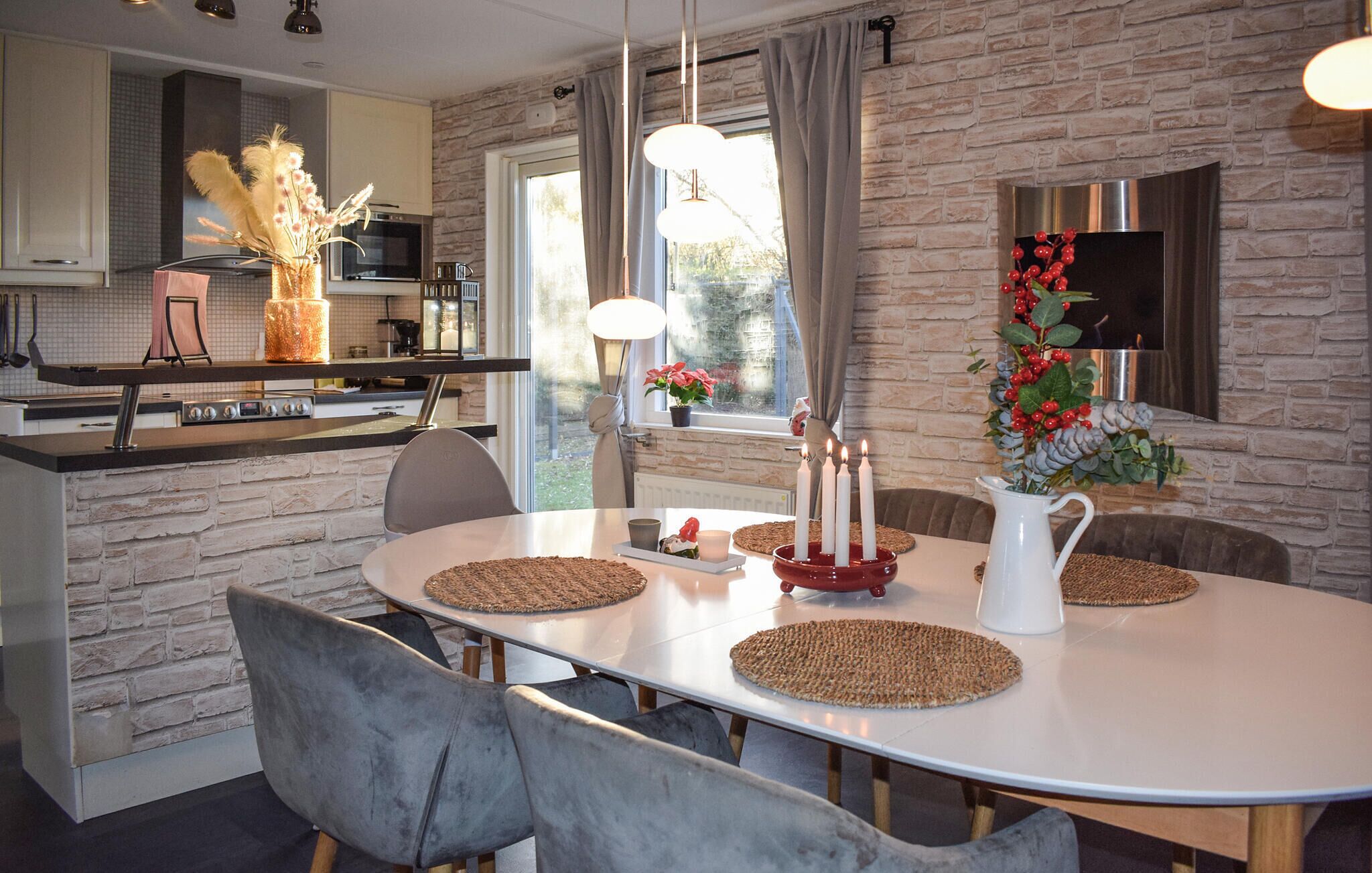 Une cuisine moderne avec un mur en pierre derrière le plan de travail, un coin repas avec une table blanche et une grande fenêtre agrémentée de rideaux.