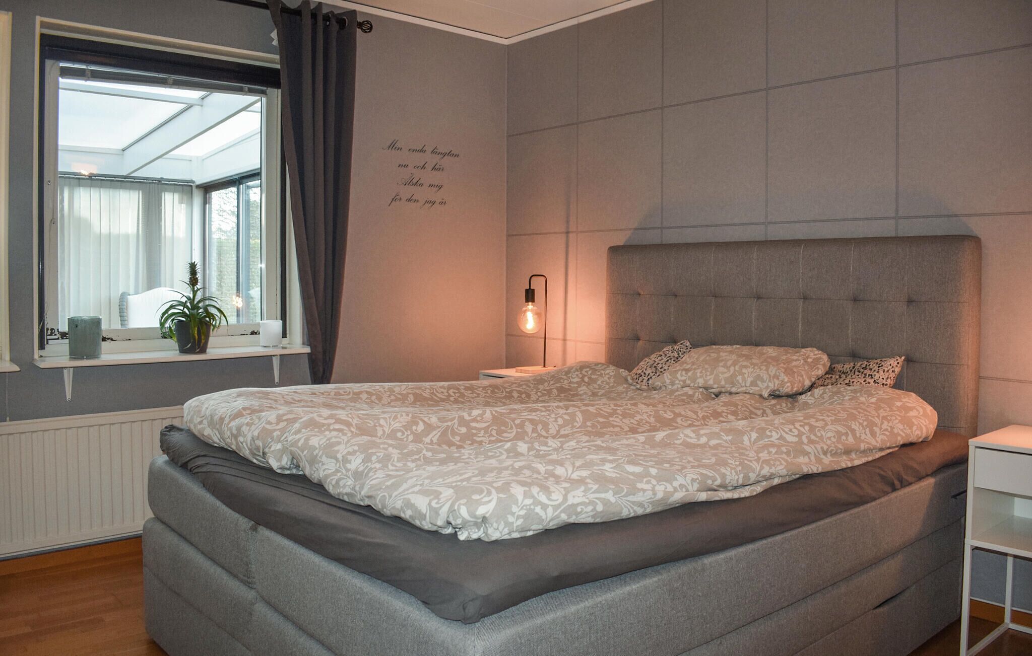 A bedroom with a large bed, a bedside table, a nightstand, a window with curtains, and a wall with a decorative text.