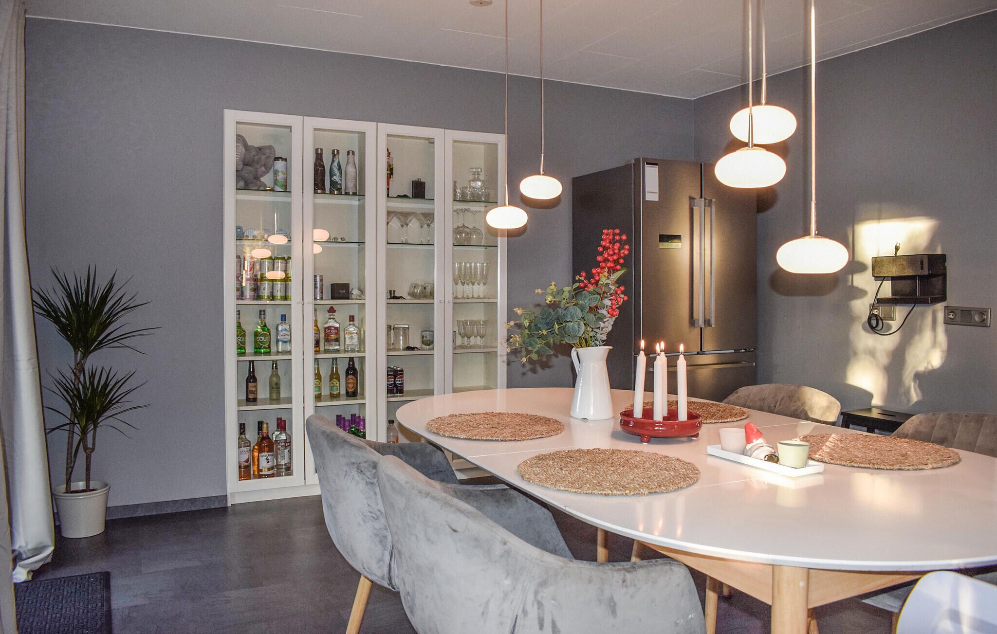 A modern dining area with a round table, grey upholstered chairs, and a cabinet with glass doors containing bottles and glasses.