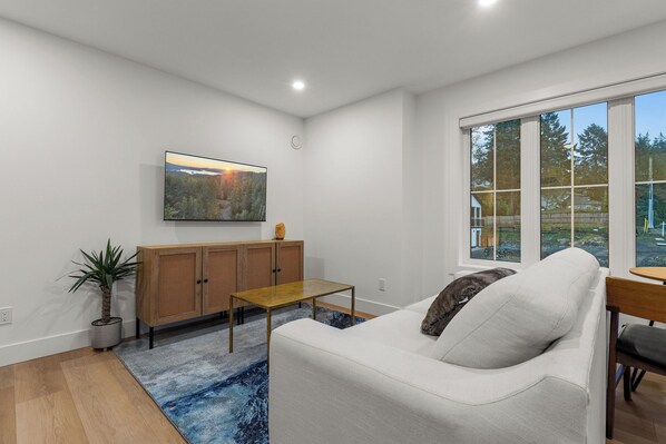 Ein modernes Wohnzimmer mit einem weißen Sofa, einem Holzkasten, einem Couchtisch und einem großen Fenster mit Blick auf Bäume und ein Haus.
