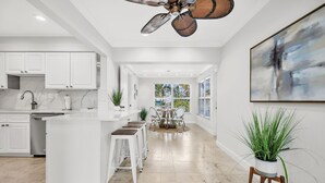 A modern kitchen with white cabinetry, a marble backsplash, and a central island with stools. There is a ceiling fan, a dining area with a table and chairs, and a large abstract painting on the wall. - Waterfront Oasis • Pool • Ocean Access. near beach.near golf.pet friendly (Pompano Beach)