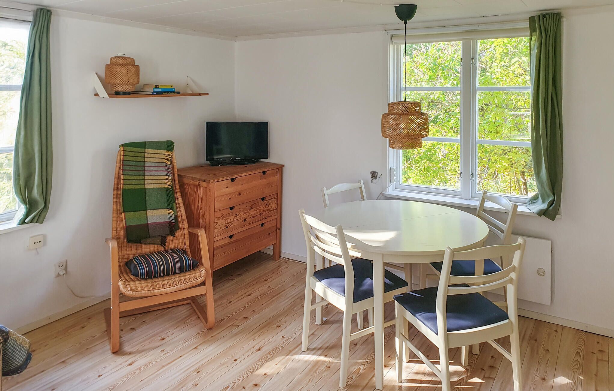 Een gezellige kamer met houten vloer, een ronde eettafel met stoelen, een televisie op een houten kast en een raam met groene gordijnen.
