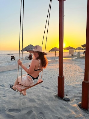 Escena de playa con un columpio, una persona balanceándose y sombrillas de playa al fondo.