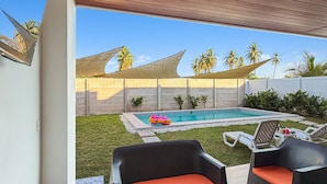 Une terrasse extérieure avec deux fauteuils en osier noir et une petite table, une piscine et une pelouse agrémentée de palmiers en arrière-plan.