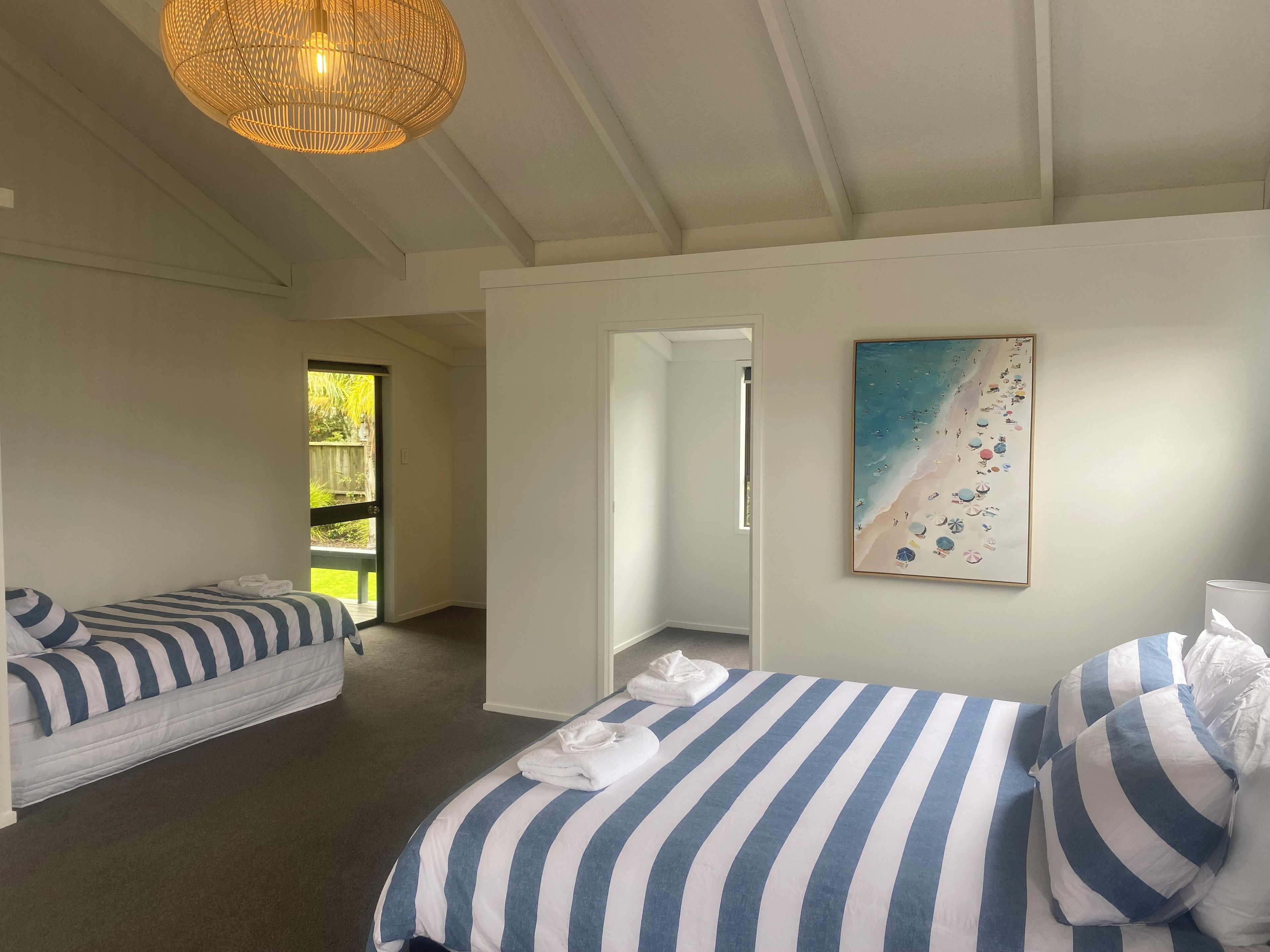 A hotel room with two beds, a painting of a beach, and a view of the outdoors.