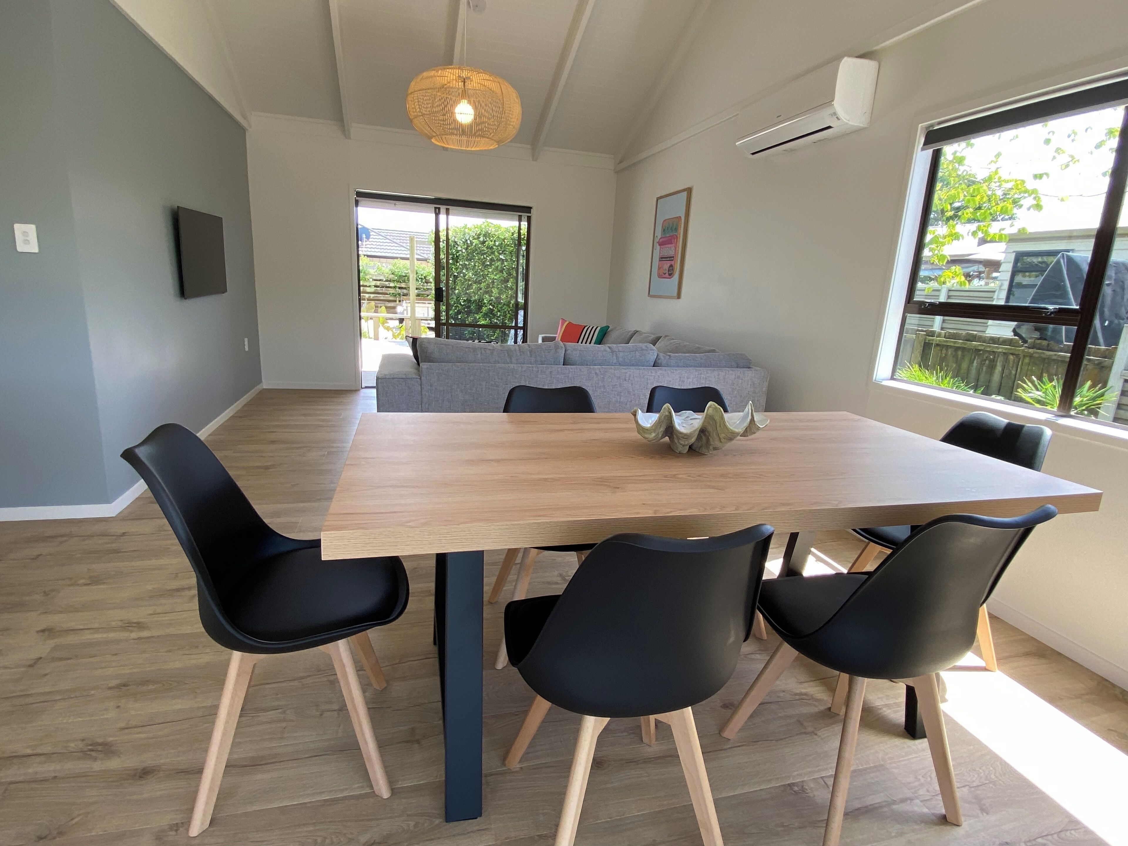 A dining area with a wooden table, black chairs, a woven pendant light, a grey sofa, and a window with a view of greenery.