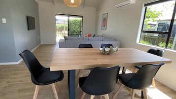 A dining area with a wooden table, black chairs, a woven pendant light, a grey sofa, and a window with a view of greenery.