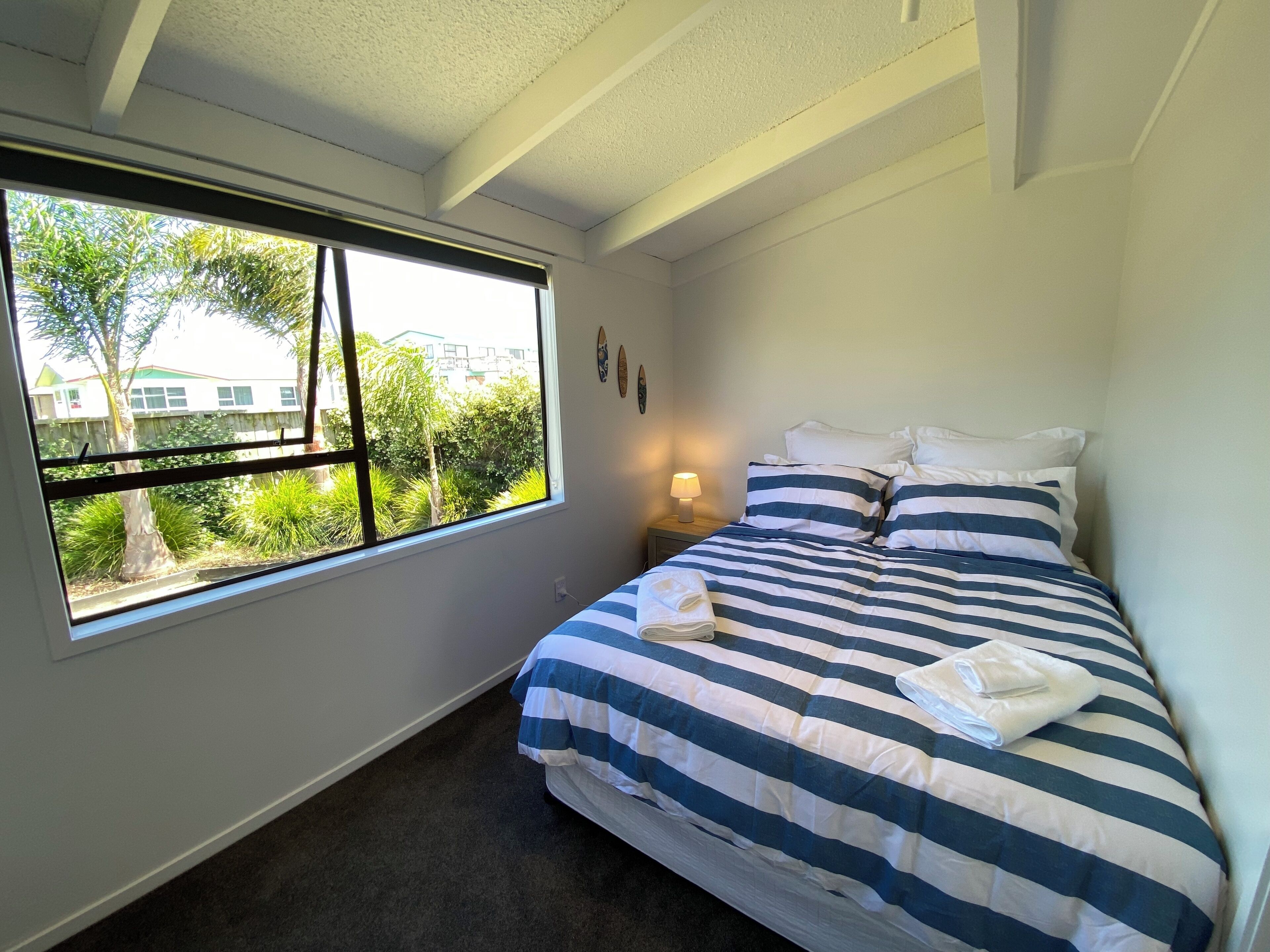 A bedroom with a large window, a bed with striped bedding, a bedside lamp, and a small wall decoration.
