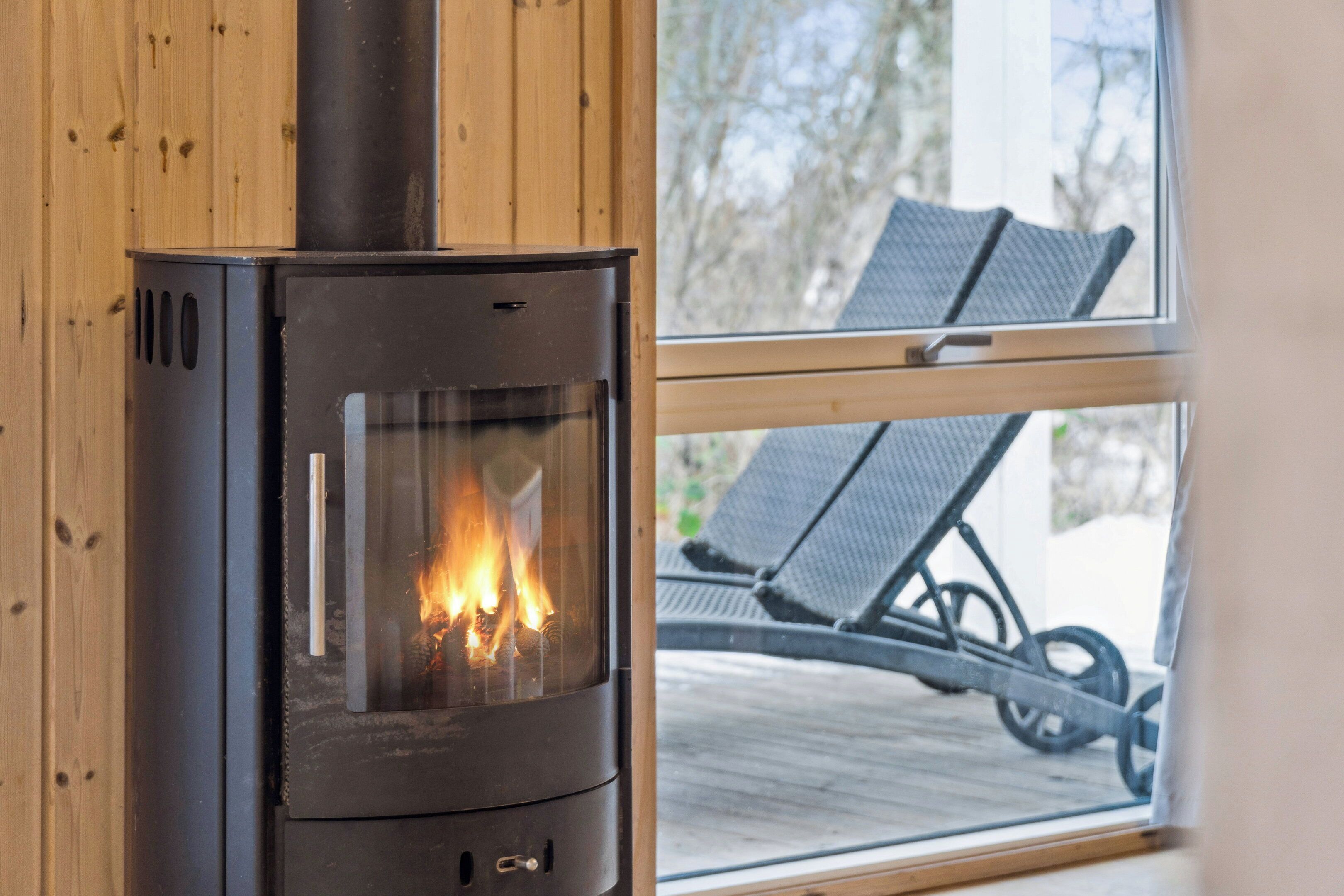 A wood-burning stove with a glass door, wooden walls, and a view of outdoor seating through a window.