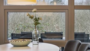 Un coin repas comprenant une table, des chaises, un saladier, un vase avec des fleurs et une fenêtre donnant sur des arbres.