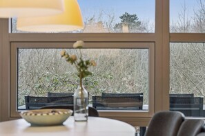 A dining area with a table, chairs, a bowl, a vase with flowers, and a window with a view of trees. - Family-friendly holiday home with spa and sauna (Knebel)