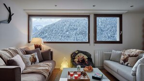 A modern living room with two sofas, a coffee table with drinks and pastries, and large windows offering a snowy mountain view.