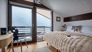 A bedroom with a large bed, a wooden desk, a chair, and a view through a sliding glass door to an outdoor balcony.