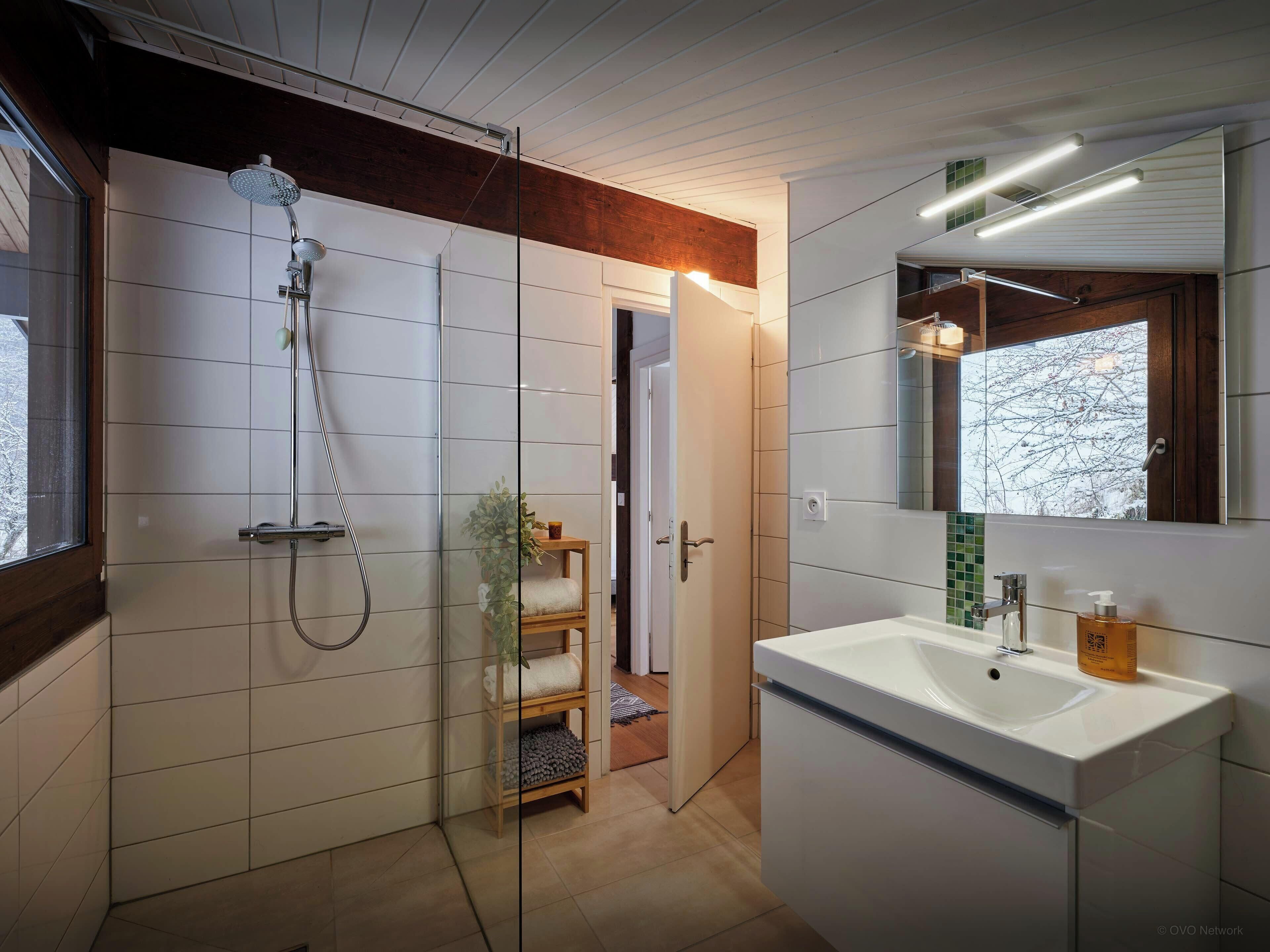 Un baño moderno con ducha acristalada, lavamanos blanco y espejo con vista a árboles.