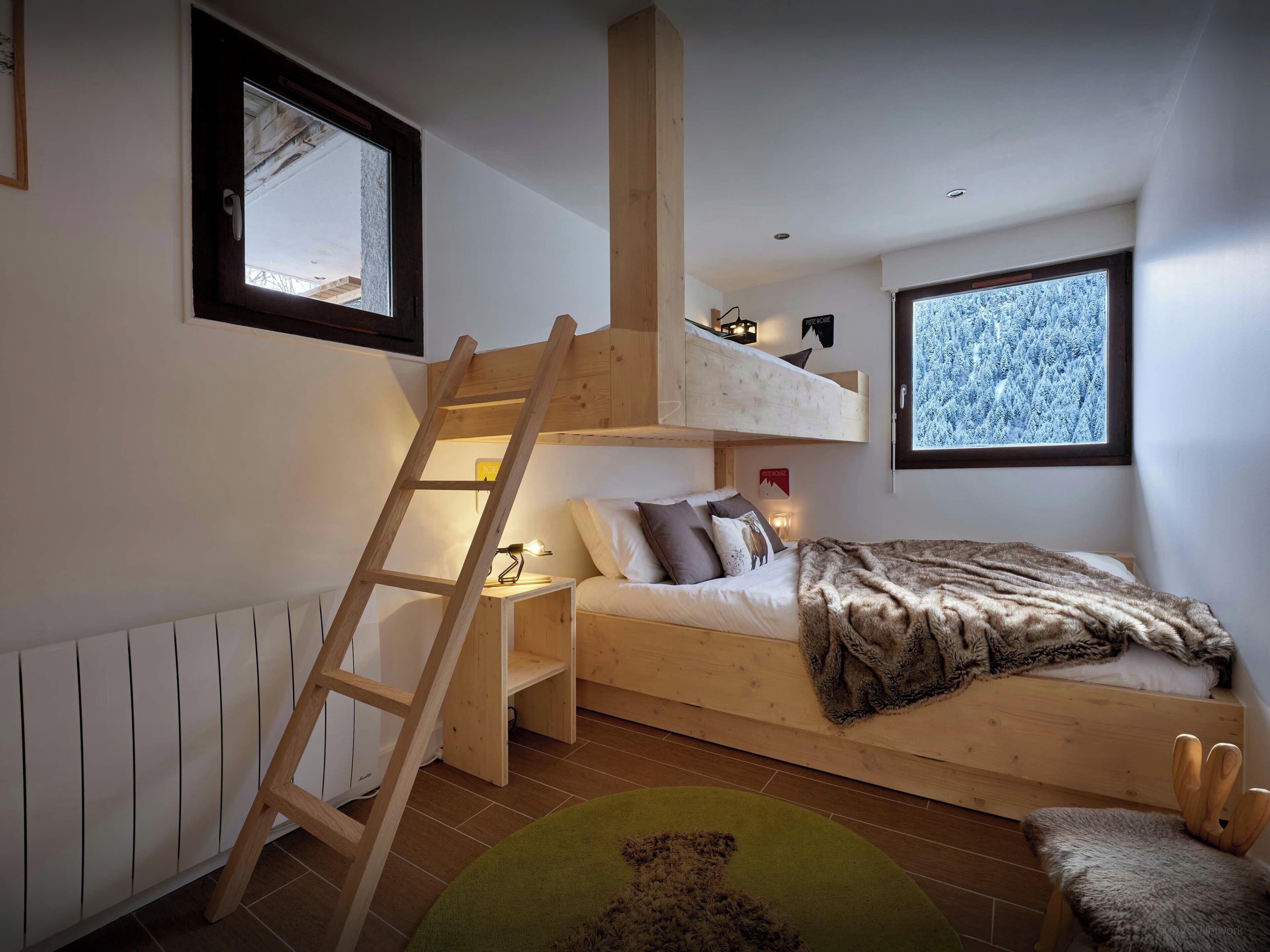 A compact bedroom with a loft bed, a wooden ladder, a green rug, and a view of a snowy landscape through a window.