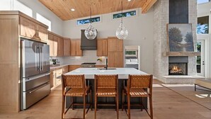 A modern kitchen with a central island, wooden cabinets, stainless steel appliances, and a stone fireplace. - NEW CONSTRUCTION 4-bedroom with INCREDIBLE VIEWS minutes to Downtown Blue Ridge! (Mineral Bluff)