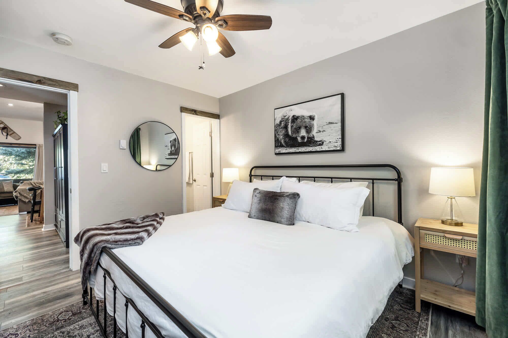A bedroom with a bed, a ceiling fan, a round mirror, a framed picture, bedside lamps, and a view of a dining area.