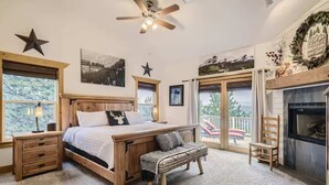 Une chambre à coucher avec un lit en bois, un banc, une table de chevet, une cheminée et de grandes fenêtres donnant sur l’extérieur.