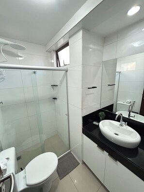 A modern bathroom with a glass shower enclosure, a white toilet, a black countertop with a sink, and a mirror. - Melhores Flats - Coração de Tambaú (João Pessoa)