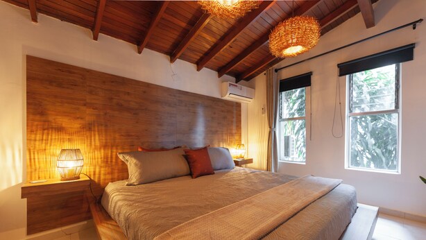 Ein Schlafzimmer mit einem Holzkopfteil, einem großen Bett mit vielen Kissen, Nachttischlampen und einem Fenster mit Blick auf Bäume.