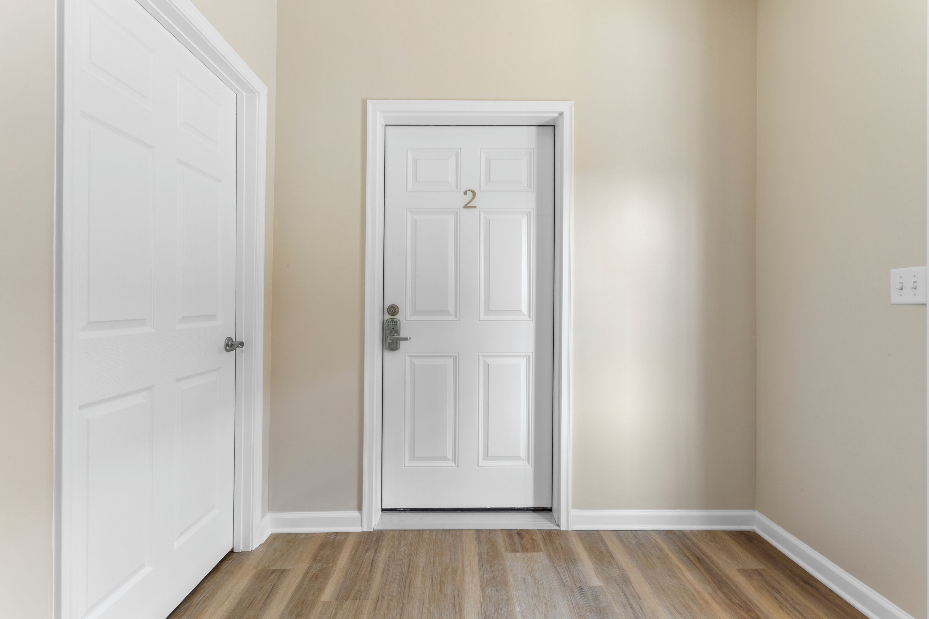 A hallway with a white door marked with the number 2, wooden flooring, and beige walls.