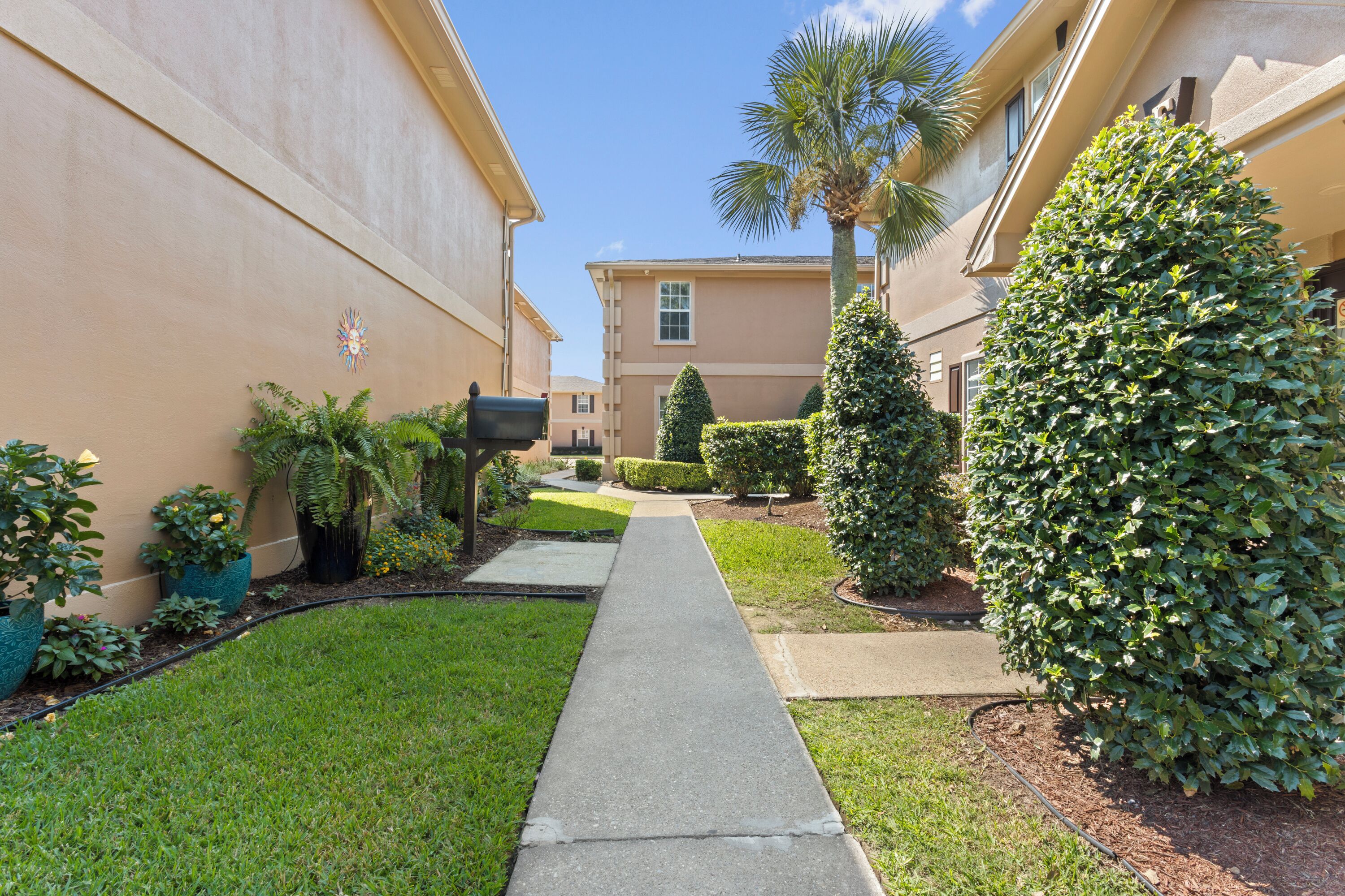 A pathway leading to a building with a beige exterior, surrounded by well-maintained grass and various shrubs.