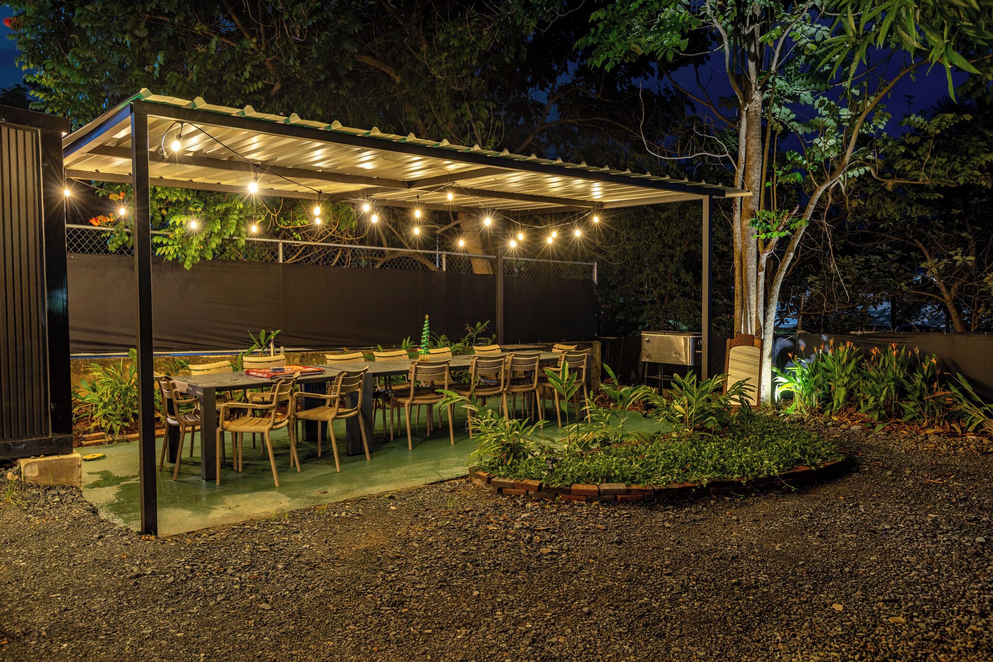 Uma área de refeições ao ar livre com varanda coberta, luzes de festa e diversas plantas.