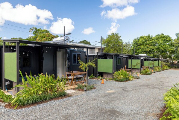 A row of modern, compact cabins with green exterior panels, a gravel driveway, and a bench in the foreground. - Tatloanis Village (Vega Baja)