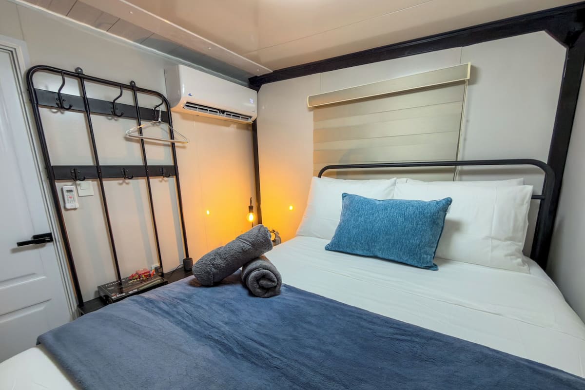 A neatly made bed with a blue pillow and a rolled towel, a bedside table with a lamp, and a wall-mounted air conditioning unit.