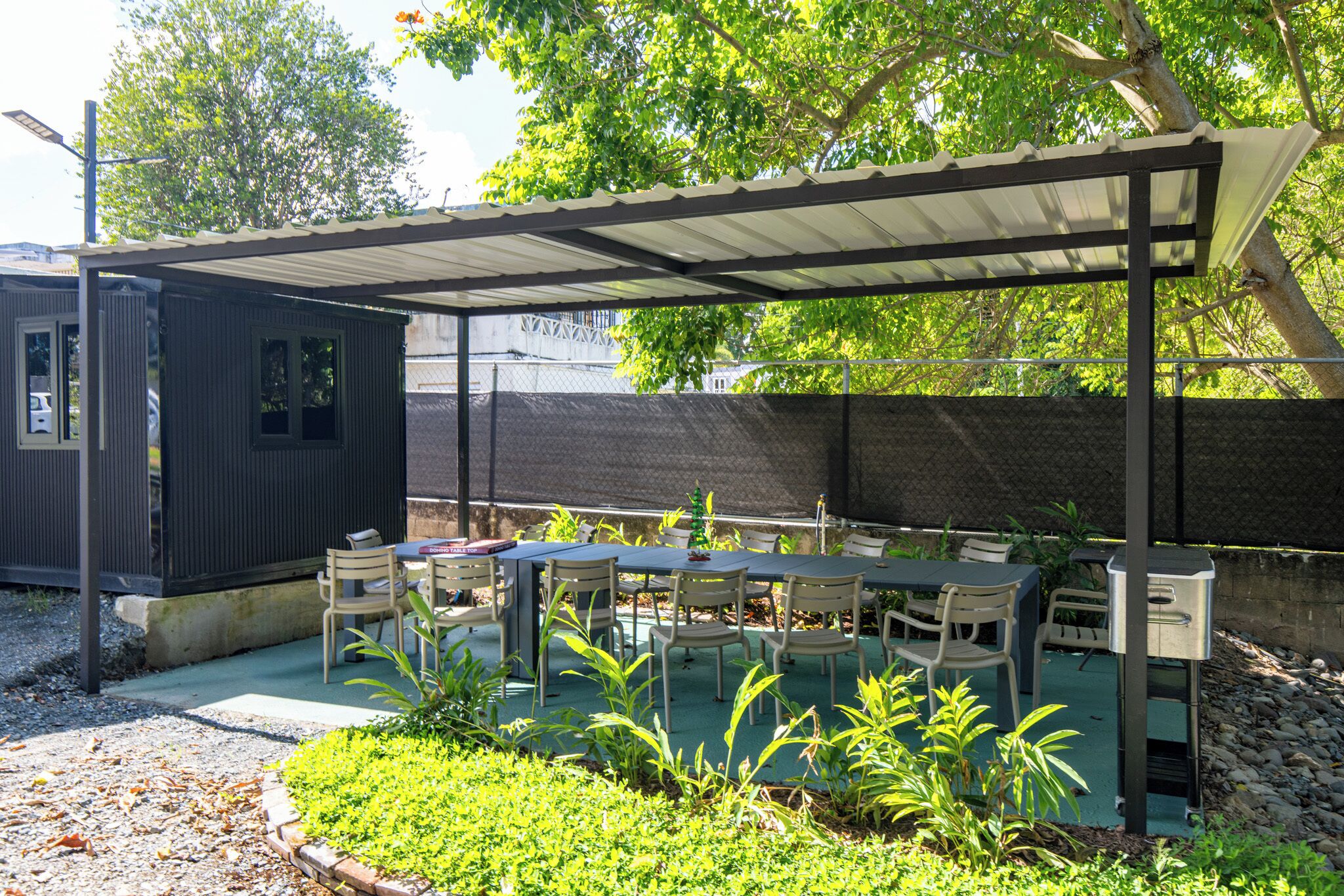 A covered outdoor patio with tables and chairs, surrounded by greenery and a fence.